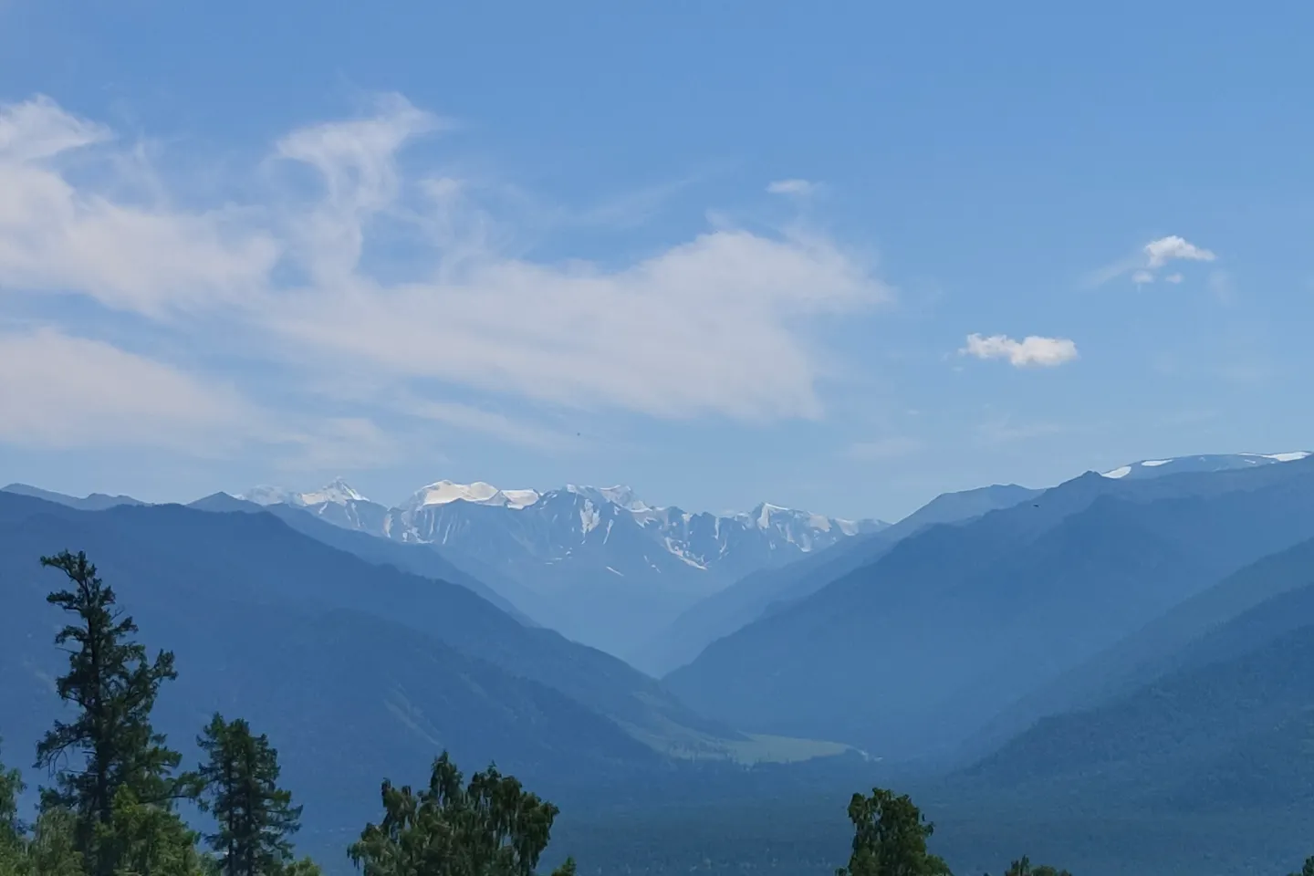 Layers of forested Altai mountains lead towards distant snow-covered peaks beneath a soft blue sky